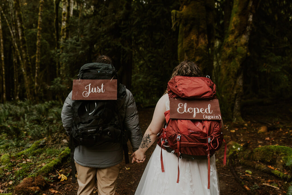 A couple in weddings outfits hiking through a mossy forest. Wearing backpacks with signs saying "just eloped"