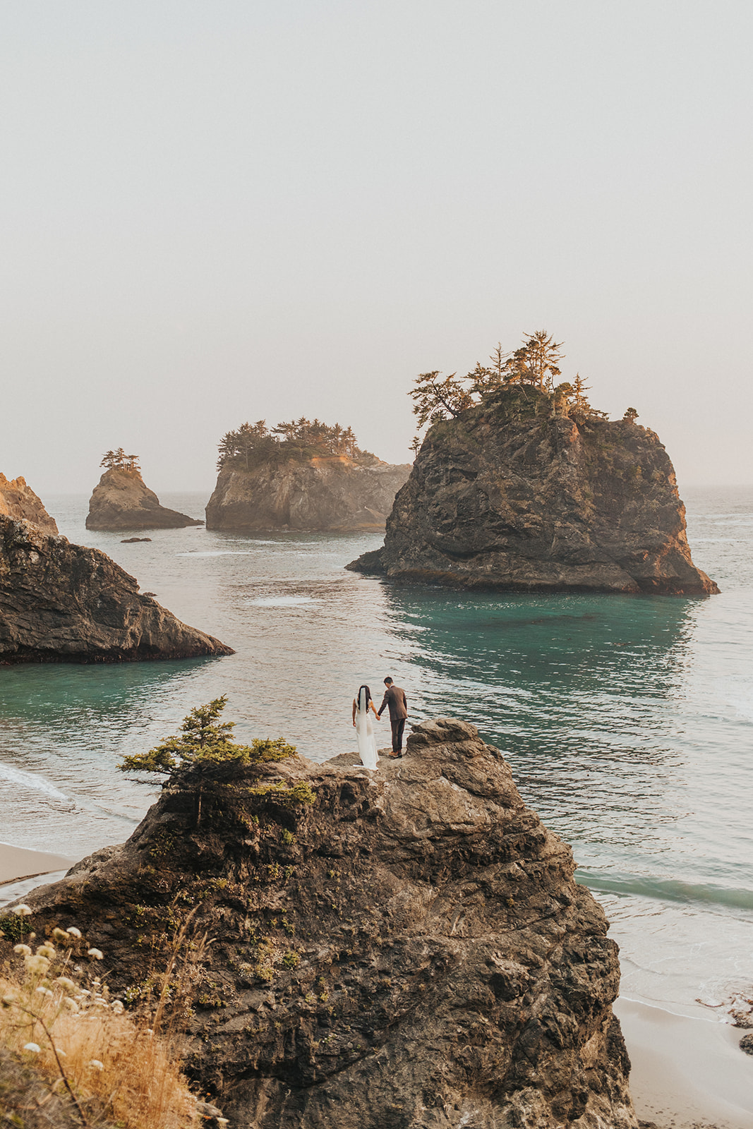 A couple in wedding attire stands on top of a rock at the edge of the ocean on the Oregon Coast with haystack rocks in the distance