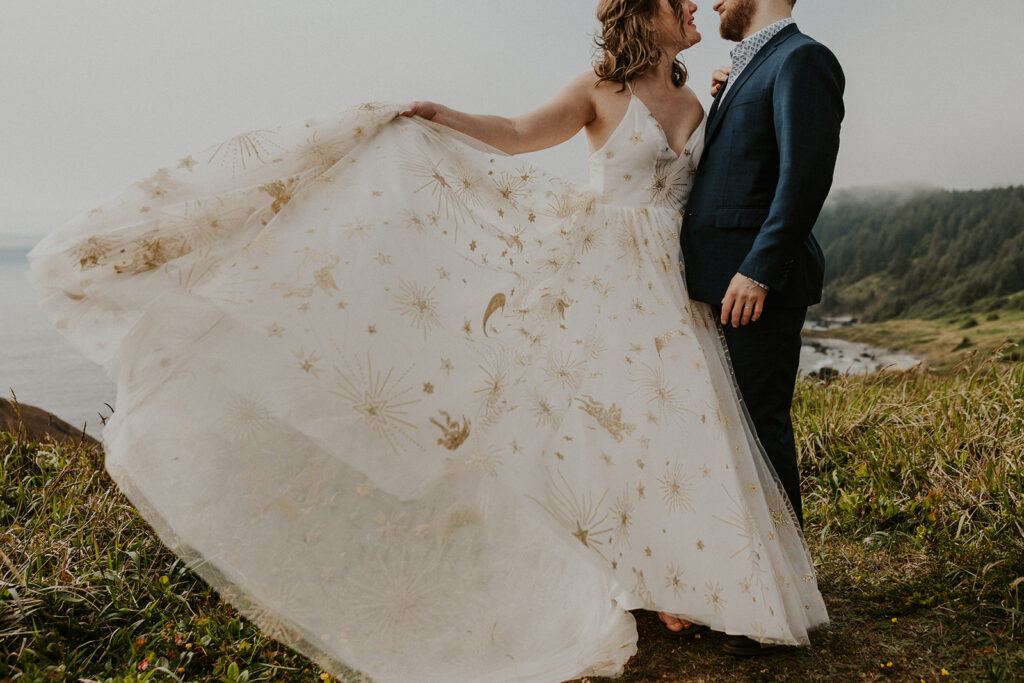 A bride and groom on an ocean bluff on the Oregon Coast, the wedding dress has stars, moons, and pegasus on it