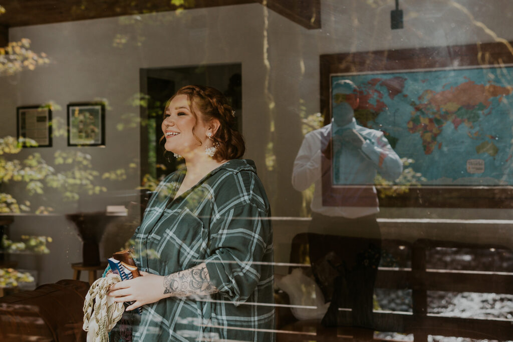 A bride looks through a window with groom reflected on the other side