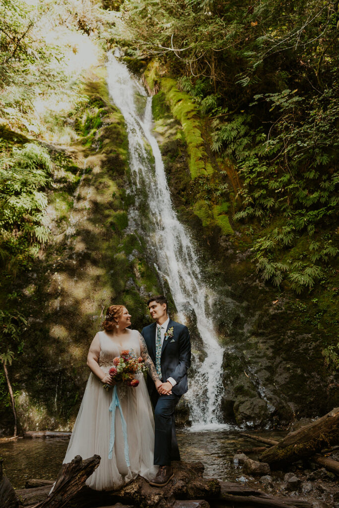 A couple in wedding attire pose by a waterfall in Olympic National Park