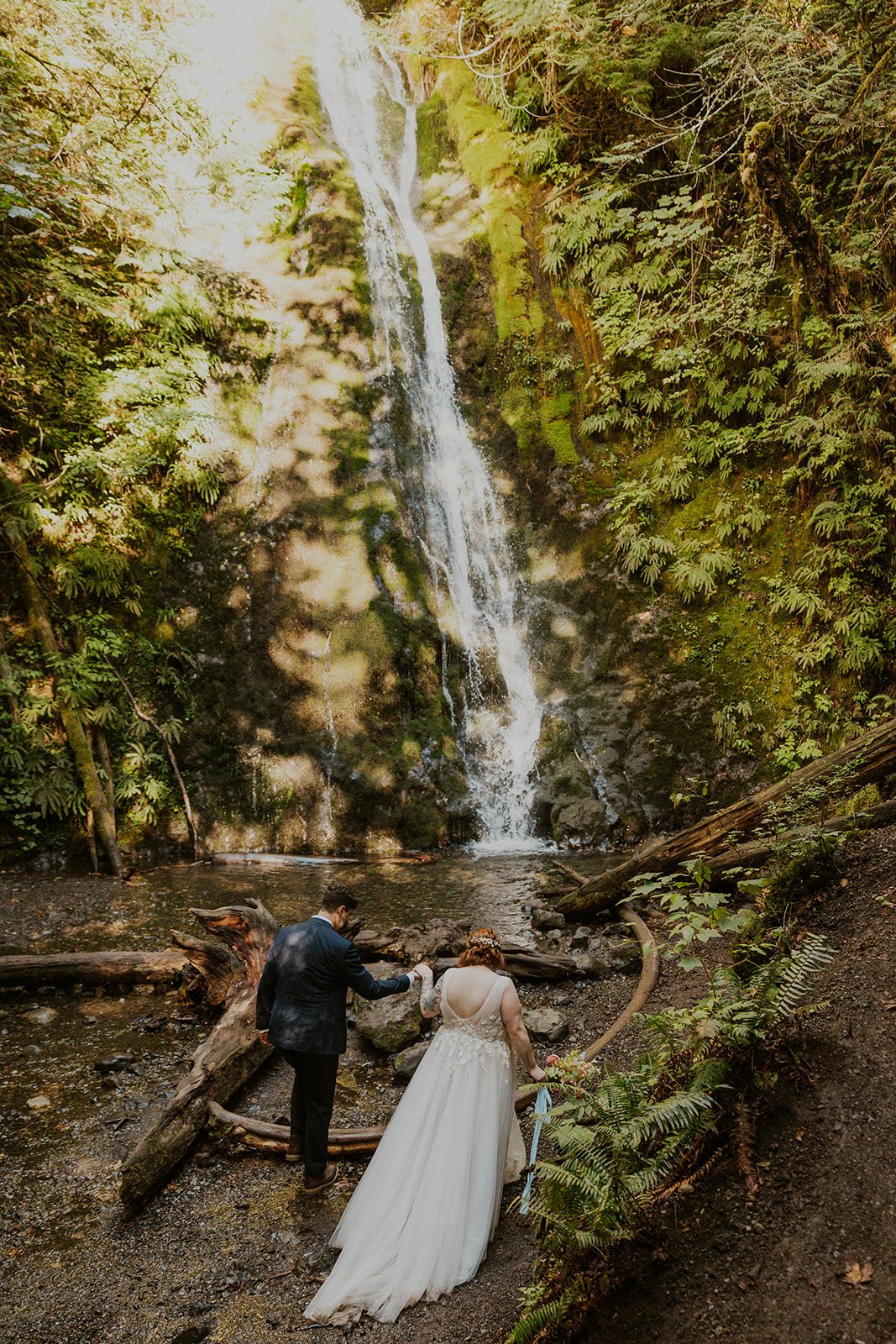 A couple in wedding attire pose by a waterfall in Olympic National Park