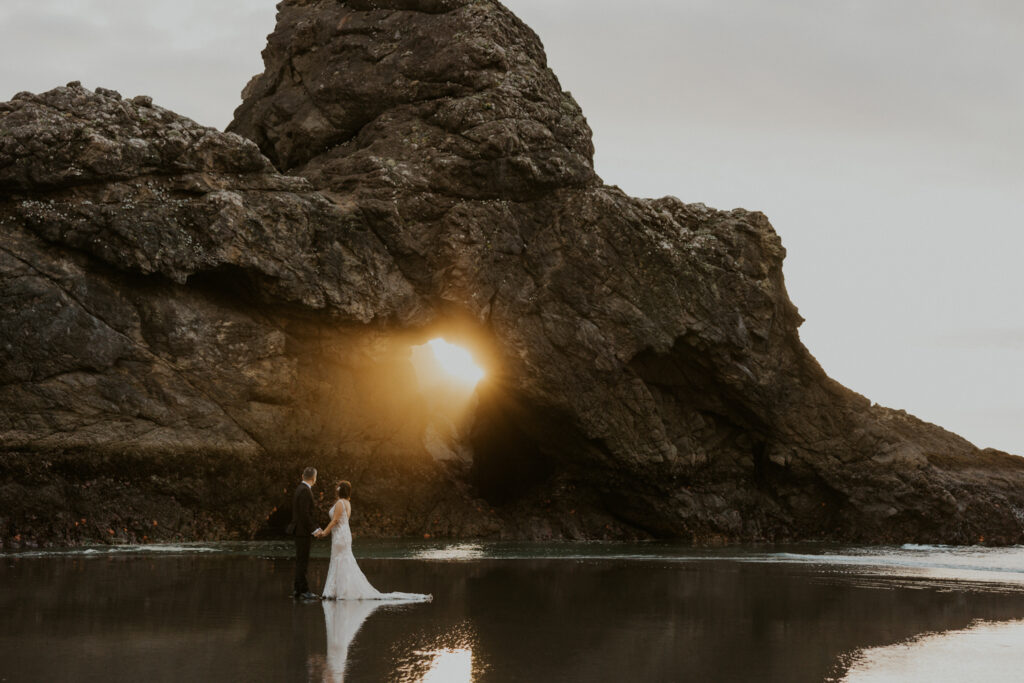 A couple in wedding attire standing under light shining through a keyhole arch on the Oregon Coast