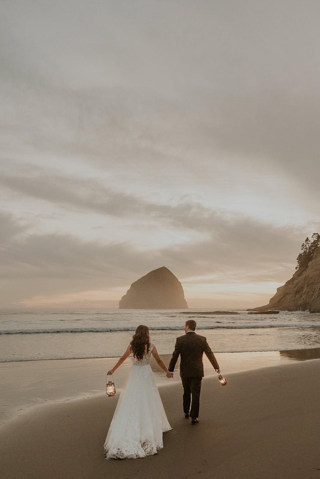A bride and groom walk on a beach in Pacific City, OR with vintage lanterns during sunset. 